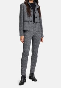 Blazer in tartan grigio con colletto alto, bordi neri e pantaloni aderenti coordinati. Tessuto liscio con una sottile texture e accenti di bottoni. Stivali neri con tacco.