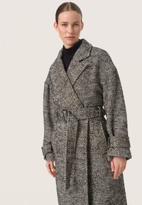 Zwart-witte houndstooth-gepatenteerde jas, gemaakt van textuurstof, met een ceintuur in de taille en brede revers, met opgerolde mouwen.