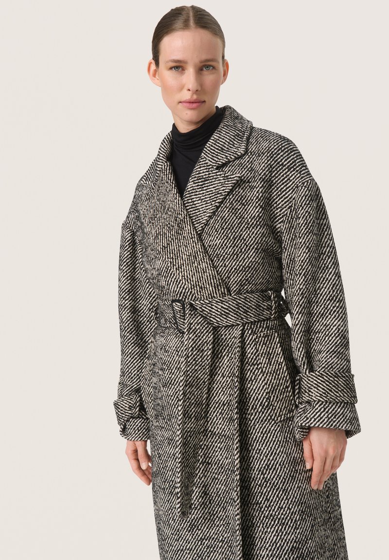 Abrigo de patrón de espigas en blanco y negro, hecho de tela texturizada, con cinturón en la cintura y solapas anchas, y puños enrollados.