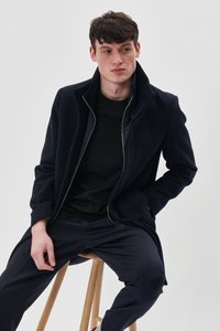 Tamsiai mėlynas vilnonis paltas su aukštu apykakle, užtrauktuku ir šoninėmis kišenėmis, dėvimas ant juodo marškinėlio ir tamsiai mėlynų kelnių, sėdintis ant medinio stalo.