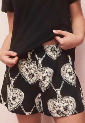 Hände heben ein schwarzes Hemd an, um schwarze Shorts mit einem weißen, herzförmigen Blumenanhängermuster zu zeigen.