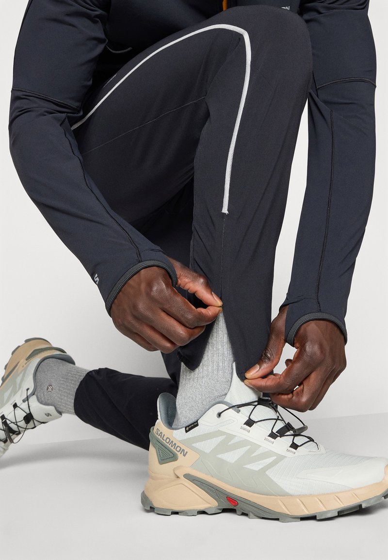 Personne ajustant la fermeture éclair sur un pantalon de sport noir près d'une chaussure de randonnée Salomon légère grise et beige sur un fond blanc.