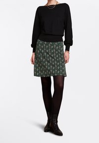 Schwarzer Pullover mit langen Ärmeln, kombiniert mit einem grünen gemusterten Rock mit geometrischem Design und schwarzen Strumpfhosen. Dazu getragen werden Knöchelstiefel.