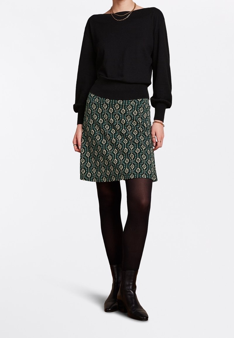 Schwarzer Pullover mit langen Ärmeln, kombiniert mit einem grünen gemusterten Rock mit geometrischem Design und schwarzen Strumpfhosen. Dazu getragen werden Knöchelstiefel.