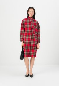 Rotes und schwarzes Karokleid mit langen Ärmeln, hohem Kragen, asymmetrischem Design und einer Brusttasche; kombiniert mit schwarzen Absatzschuhen und einer Handtasche.
