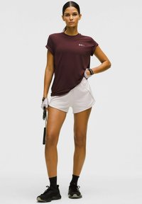 Sportos nő áll, ugrókötéllel a kezében, bordó T-shirt-ben, fehér rövidnadrágban, fekete zokniban, fekete edzőcipőben és fehér edzőkesztyűben.