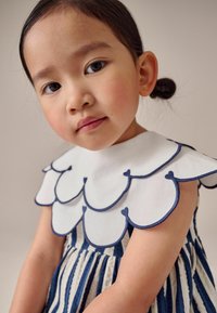 Kinderkleid mit einem weißen Kragen, der navy-blauem gewellten Rand verziert ist. Das Kleid hat vertikale Streifen in navy-blau und weiß mit einer gerafften Taille.