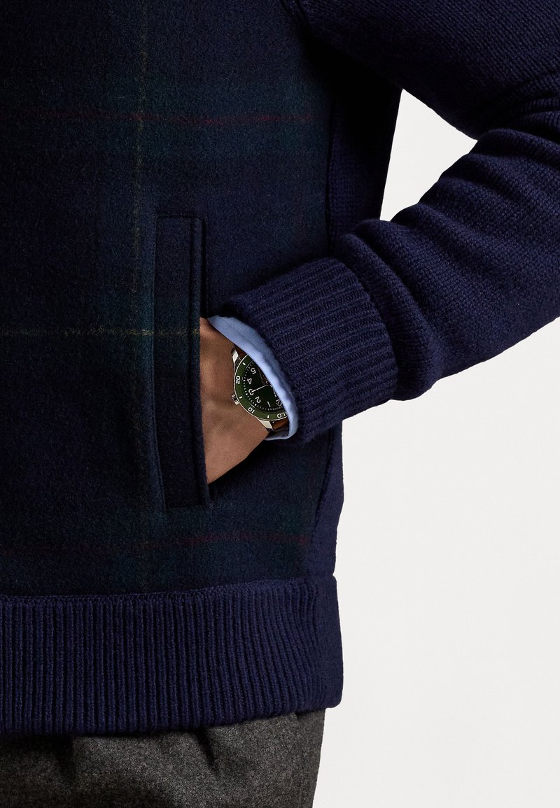 Navy knitted sweater with a textured finish, featuring a side pocket and ribbed cuffs. A watch with a black dial is visible on a hand.