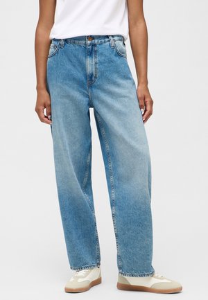 Person trägt hellblaue Jeans mit weitem Bein, weiße Sneakers mit beigen Akzenten und ein in die Hose gestecktes weißes Hemd, steht vor einem schlichten Hintergrund.