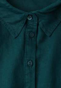 Chemise vert foncé à col avec trois boutons visibles fermés, montrant un tissu texturé et des coutures autour du col.