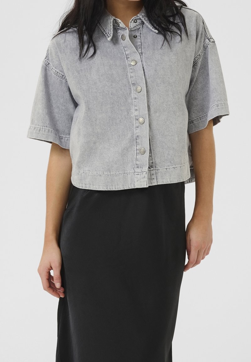 Femme portant une chemise en jean gris clair raccourcie, à boutons et manches courtes, ainsi qu'une jupe noire taille haute sur un fond uni.