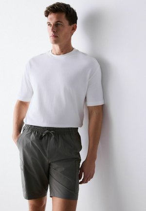 Uomo con indosso una camicia bianca a maniche corte e pantaloncini grigi con coulisse, in piedi contro un muro bianco, che guarda di lato.