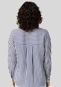 Maglietta a righe blu navy e bianca a maniche lunghe con schiena plissettata, dotata di orlo curvo e polsini abbottonati. Il tessuto sembra leggero e strutturato.