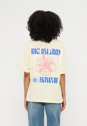 Person med krøllet hår iført en cremefarvet T-shirt med en lyserød hibiscusblomst og blå tekst, der lyder Big Island Hawaii, kombineret med blå jeans.