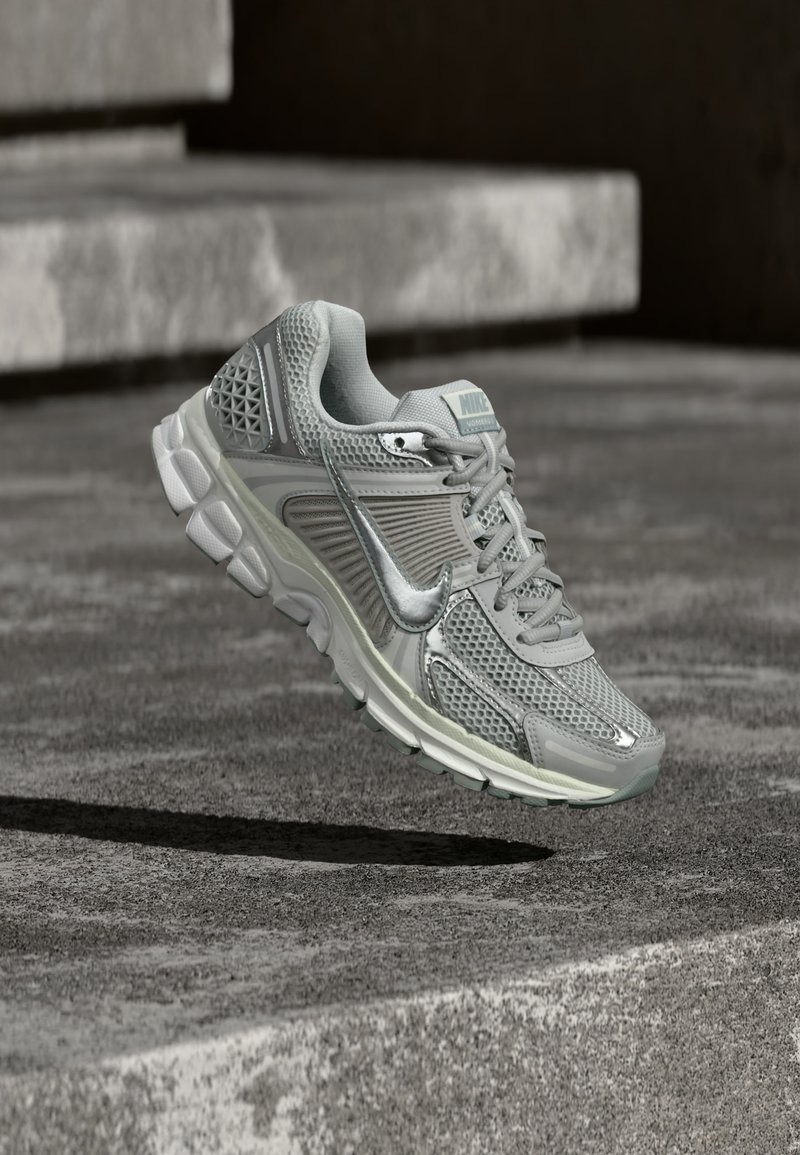 Pilkas Nike bėgimo batelis su tinklelio viršumi ir paminkštinta padais, sklandantis virš betoninių laiptų.