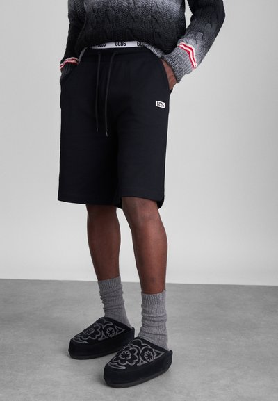 Black cotton shorts with an elastic waistband, side pockets, logo patch, paired with gray ribbed socks and black patterned slip-on shoes.