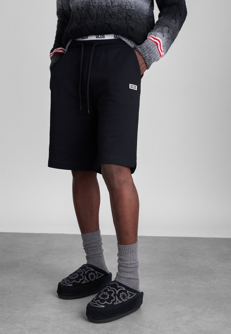 Schwarze Baumwollshorts mit einem elastischen Bund, seitlichen Taschen, einem Logo-Patch, kombiniert mit grauen gerippten Socken und schwarzen gemusterten Slippers.