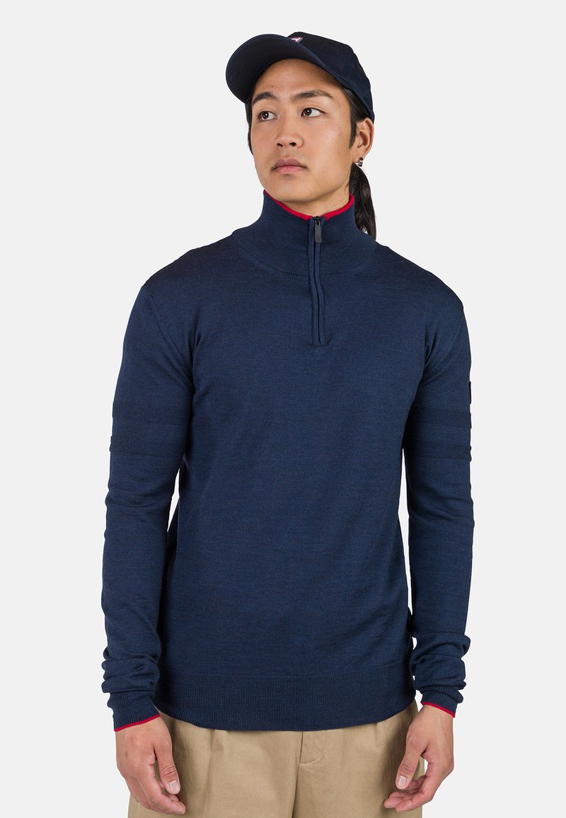 Navy-blauer Strickpullover mit hohem Kragen und einem Viertel-Reißverschluss. Mit roten Akzenten am Kragen und an den Bündchen. Lange Ärmel mit strukturiertem Detail.