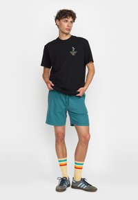 Camiseta de algodón negra con logotipo frontal, pantalones cortos en color teal, calcetines amarillos con rayas de colores del arcoíris y zapatillas grises con acentos azules.