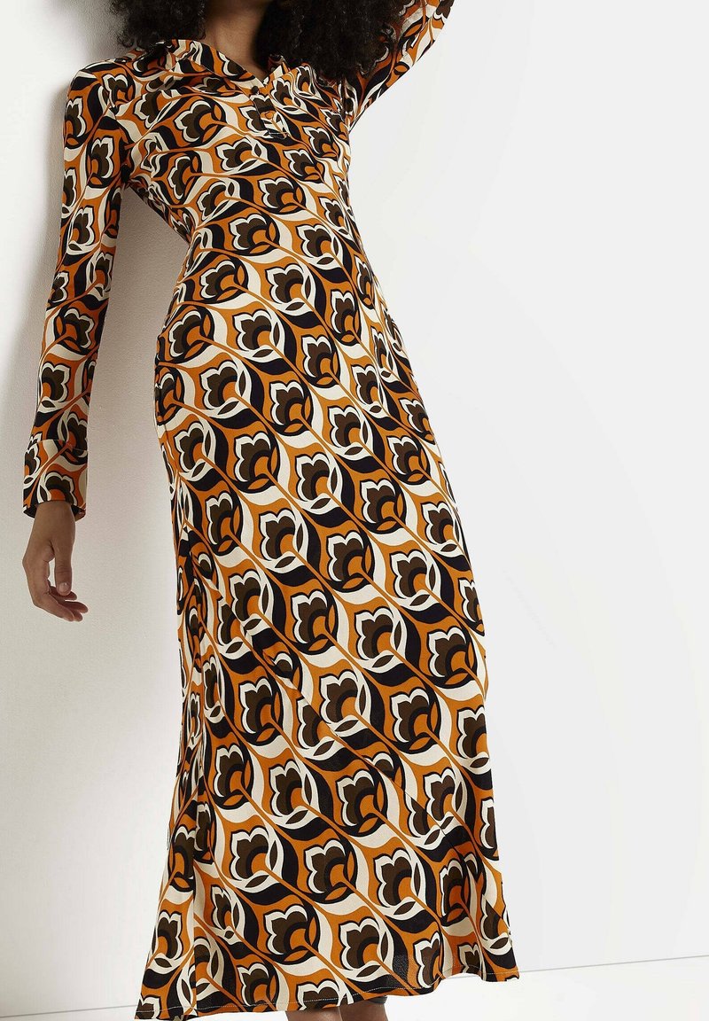 Femme portant une robe longue à manches longues avec un motif floral géométrique audacieux orange, noir et crème sur un fond blanc uni.