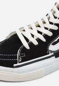 Svart semsket høyhælte sneakers med hvite snøringer, kontrasterende sømmer og gummisåle, med et dekorativt design på siden.