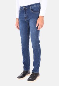 Jeans in denim blu con una vestibilità slim, caratterizzati da una chiusura a bottone, tasche frontali e un leggero sbiadimento sulle cosce. Indossati con scarpe nere.