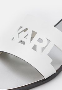 Sandal in pelle bianca con lettere ritagliate che formano "KART", dotato di suola piatta e una soletta liscia in argento. Design minimalista e forma snella.