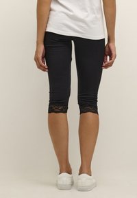 Leggings noirs avec une bordure en dentelle à l'ourlet, longueur cheville, fabriqués en tissu extensible, associés à des baskets blanches et un haut blanc ample.
