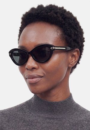 Femme aux cheveux courts et bouclés portant des lunettes de soleil noires œil de chat et un pull gris à col roulé côtelé, regardant légèrement sur le côté.