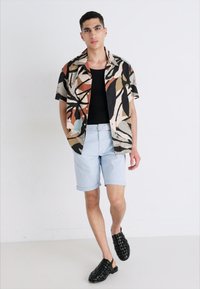 Chemise à manches courtes à motifs dans des tons beige, noir et vert, portée sur un débardeur noir ; short bleu clair et sandales tressées noires.