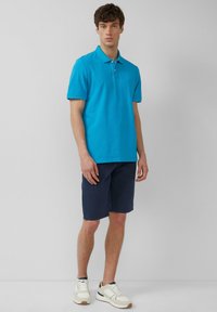 Türkisfarbenes Poloshirt aus Baumwollstoff, mit kurzen Ärmeln und Kragen. Kombiniert mit marineblauen Shorts und weißen Sneakern mit dunklen Akzenten.