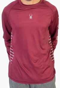 Bordowa sportowa koszulka z długim rękawem z logo pająka na klatce piersiowej i cienkimi białymi paskami po bokach. Wykonana z gładkiego, lekkiego materiału.