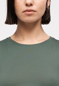 Γυναίκα που φοράει πράσινο μπλουζάκι με στρογγυλή λαιμόκοψη, με κοντά σκούρα μαλλιά, που δείχνει το πρόσωπό της από τη μύτη και κάτω καθώς και μέρος του λαιμού και των ώμων της.