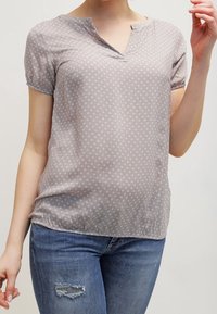 Blusa grigio chiaro a maniche corte con scollo a V, caratterizzata da un delicato motivo a pois e una vestibilità leggermente ampia. Tessuto morbido e orlo arricciato.