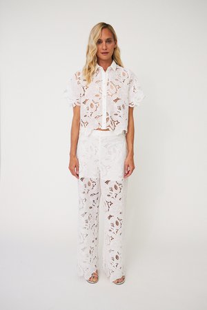 Γυναίκα με μακριά ξανθά μαλλιά φοράει λευκό δαντελένιο κοντομάνικο τοπ με κουμπιά και αντίστοιχο φαρδύ παντελόνι δαντέλας, στέκεται μπροστά σε απλό φόντο.