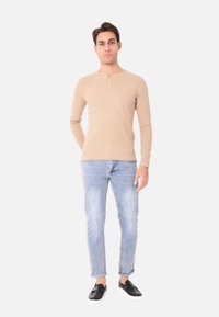 Maglietta henley beige a maniche lunghe con dettaglio di bottoni, abbinata a jeans blu chiaro e scarpe slip-on nere. Design semplice e aderente.