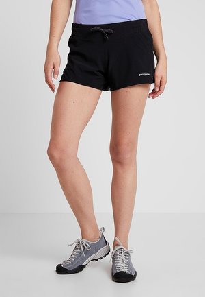 Kvinde iført sorte Patagonia shorts med snørelukning og grå vandresko, stående på en grå gulv mod en lysegrå baggrund.