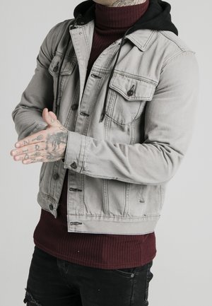 Homme portant une veste en denim gris clair par-dessus un sweat à capuche noir et un pull à col roulé bordeaux, montrant des mains tatouées jointes devant.
