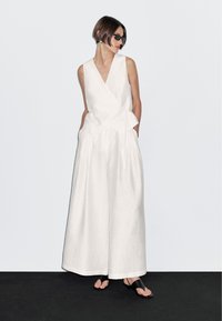 Kobieta z krótkimi brązowymi włosami, nosząca czarne okulary przeciwsłoneczne, bezrękawowy biały top, szerokie białe spodnie i czarne sandały na tle białego tła.