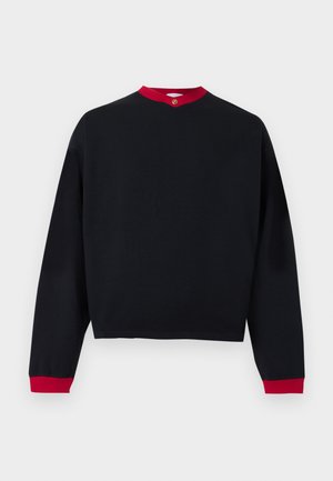 Férfi áll egy szürke falnak támaszkodva, fekete pulóvert visel piros ing fölött, kék farmert, fekete cipőt és arany medálos nyakláncot.