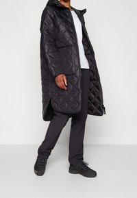 Abrigo largo acolchado negro con capucha, que presenta un cierre frontal, aberturas laterales y que se combina con pantalones oscuros y zapatos negros texturizados.