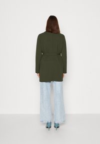 Abrigo largo verde oliva con cintura ceñida, silueta recta y mangas largas, combinado con vaqueros de pierna ancha en azul claro y tacones en tono teal.