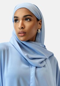 Jasnoniebieski hijab wykonany z gładkiego materiału, układający się wokół twarzy w delikatne fałdy. Modelka nosi także pasujący strój, prezentujący prosty krój.