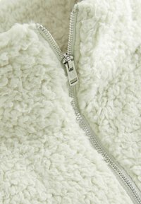 Gros plan d'une veste polaire de couleur claire avec une fermeture éclair argentée partiellement fermée et un tissu texturé et doux.