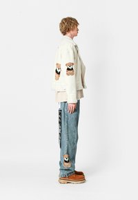 Chaqueta blanca con bordado de oso en las mangas, combinada con jeans de color azul claro que presentan un detalle de oso. Calzado marrón. Silueta de corte recto.