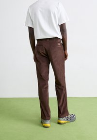 Hnědé kordové kalhoty se střihem rovného nohavice, s zadními kapsami a malým logem. Nosí se s bílou košilí s krátkým rukávem.