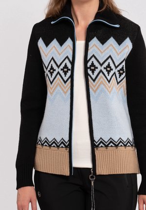 Vrouw draagt een vest met rits en zwarte mouwen, lichtblauw en beige zigzagpatroon, over een wit shirt en zwarte broek.