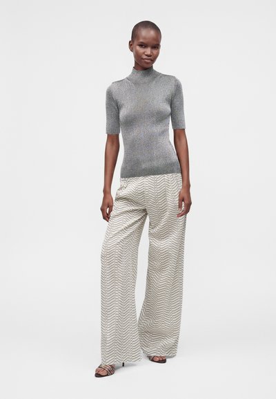 Femme portant un col roulé gris à manches courtes et un pantalon blanc à larges jambes et motifs, debout devant un fond clair uni.