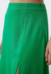 Jupe verte avec une texture lisse, dotée d'une fente sur le côté et d'une fermeture éclair discrète à l'arrière. Le tissu a un léger éclat.