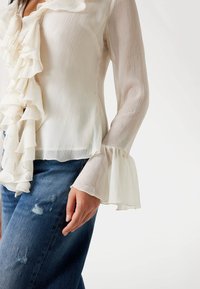 Bluză albă, cu volane, cu mâneci lungi în formă de clopot; fabricată dintr-un material ușor, texturat. Asortată cu blugi albaștri, deteriorați.